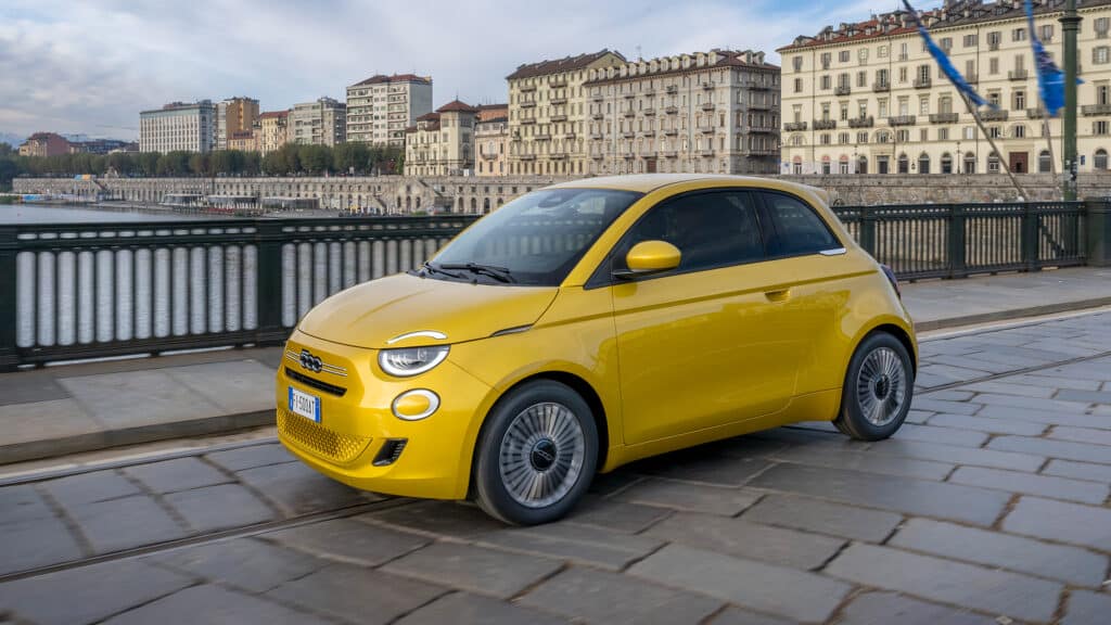 Auf diesem Bild ist ein leuchtend gelber Fiat 500e zu sehen, der auf einer gepflasterten Straße durch eine europäische Stadt mit historischen Gebäuden und einem Fluss im Hintergrund fährt.