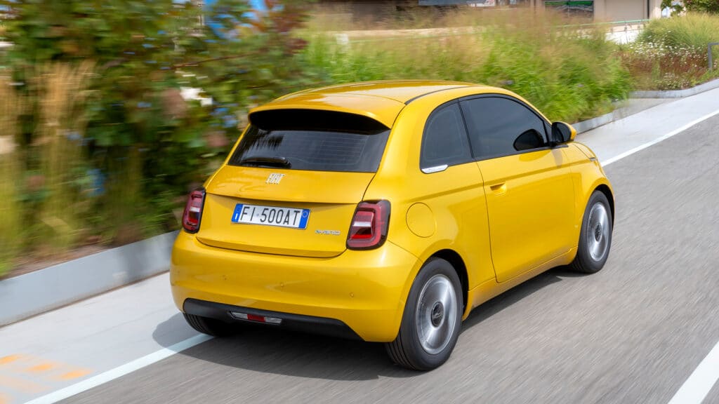 Auf diesem Bild ist ein gelber Fiat 500 Hybrid von hinten auf einer Straße zu sehen, wobei der Hintergrund durch die Bewegung unscharf dargestellt ist.