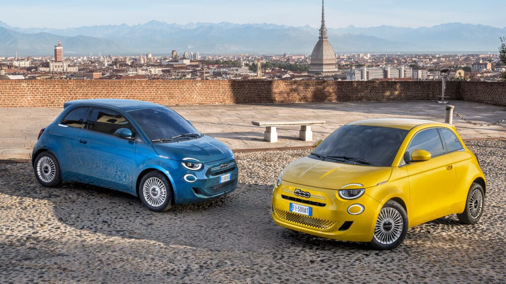 Auf dem Bild sind zwei Fiat 500 Elektroautos in Blau und Gelb auf einem gepflasterten Platz abgebildet, mit der Skyline von Turin und den Alpen im Hintergrund.