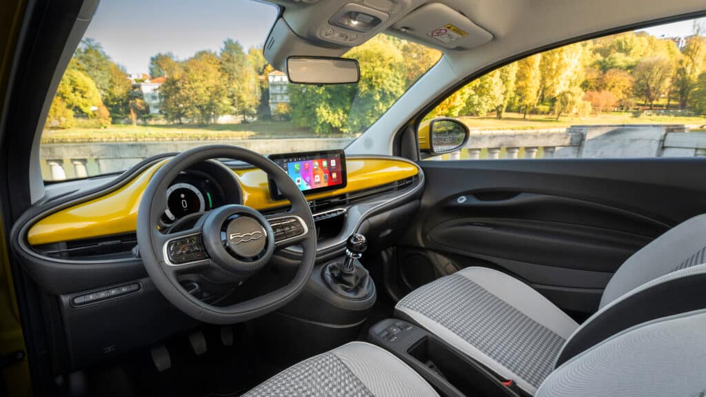 Das Bild zeigt den Innenraum eines Fiat 500 mit einem markanten gelben Armaturenbrett und dem Blick durch die Windschutzscheibe auf eine herbstliche Landschaft.