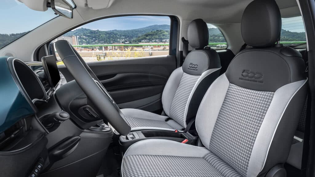 Das Bild zeigt den Innenraum eines Fiat 500, insbesondere den Beifahrersitz mit seinem markanten '500' Logo, das Lenkrad und eine weite Aussicht auf Berge und Gebäude durch das Seitenfenster.