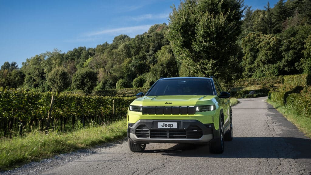 New Jeep Compass SUV Exterior: Frontalansicht in Hellgrün während der Fahrt auf einer von Bäumen gesäumten Straße.