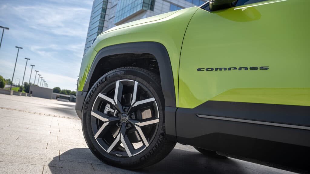 Auf diesem Bild ist die Vorderseite eines leuchtend grünen Jeep Compass mit markanten Felgen und Michelin-Reifen auf einem gepflasterten Platz vor einer modernen Gebäudekulisse zu sehen.