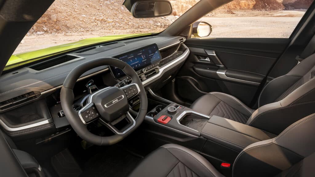 Das Bild zeigt den modernen Innenraum eines Jeeps Compass mit Lenkrad, digitalen Displays, einer detailreichen Mittelkonsole und zweifarbigen Sitzen, während durch die Windschutzscheibe eine staubige Offroad-Landschaft zu erkennen ist.