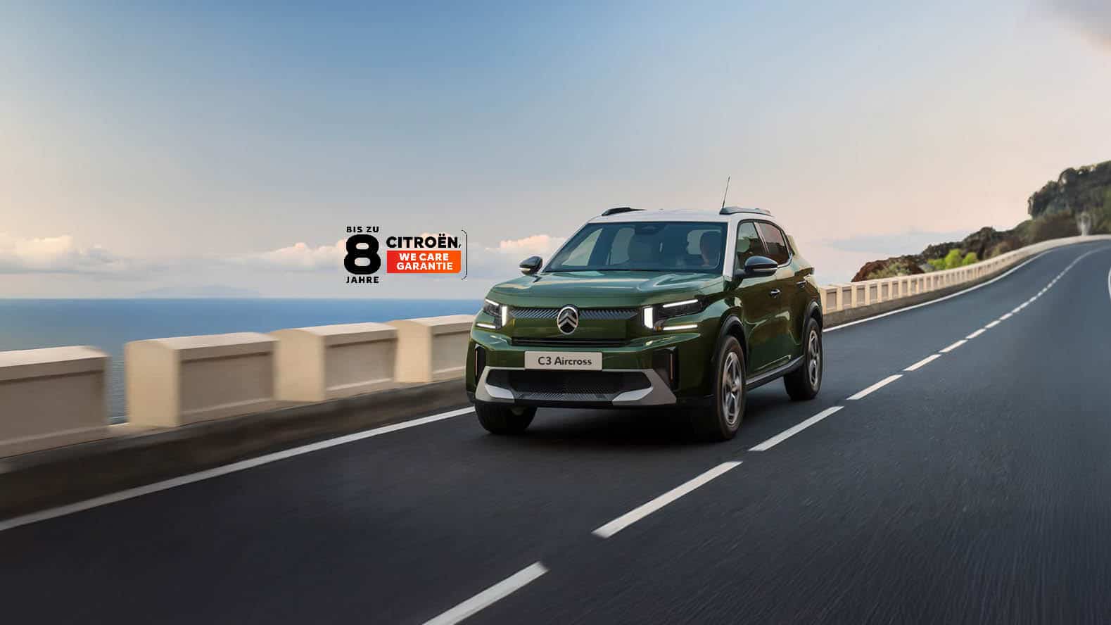 Auf diesem Bild ist ein grüner Citroën ë-C3 Aircross auf einer Küstenstraße zu sehen, mit dem Meer im Hintergrund und einer Werbeaufschrift für die „Bis zu 8 Jahre Citroën We Care Garantie“.