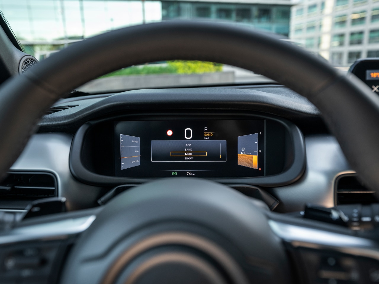 Auf diesem Bild ist das Interieur eines Autos zu sehen, mit einem Lenkrad im Vordergrund und einem digitalen Armaturenbrett, das den Fahrmodus 'SAND', die Geschwindigkeit von 0 km/h und eine Restreichweite von 160 km anzeigt.