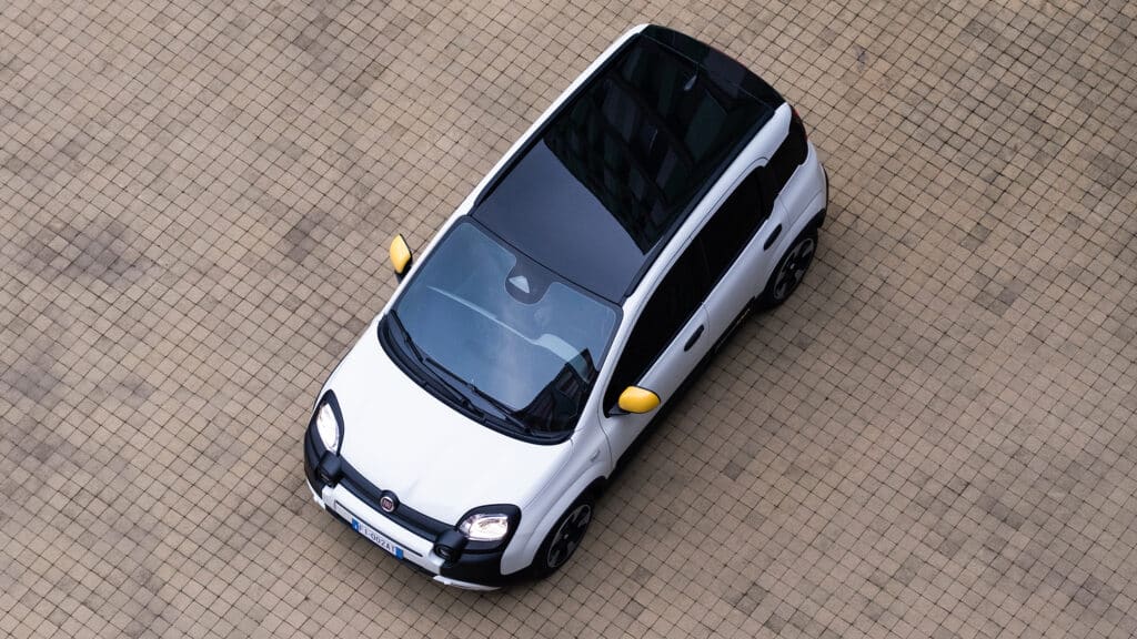 Auf diesem Bild ist ein weißer Fiat Pandina mit schwarzem Dach und gelben Seitenspiegeln aus der Vogelperspektive auf einem gepflasterten Boden zu sehen.