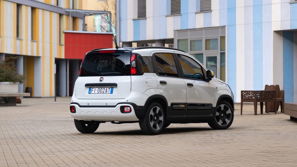 Auf diesem Bild ist ein weißer Fiat Pandina auf einem modernen Platz mit gestreiften Gebäuden im Hintergrund zu sehen.