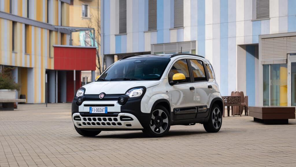 Ein weißer Fiat Pandina mit gelben Spiegeln steht auf einem gepflasterten Platz vor modernen Gebäuden mit farbigen Streifen.