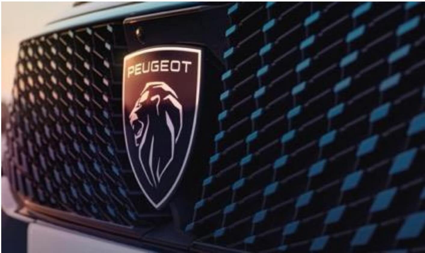 Das Bild zeigt die Front eines modernen Autos mit dem Peugeot-Logo, das einen stilisierten Löwenkopf und den Schriftzug 'PEUGEOT' darstellt, vor einem dunklen Kühlergrill mit blauen Akzenten.
