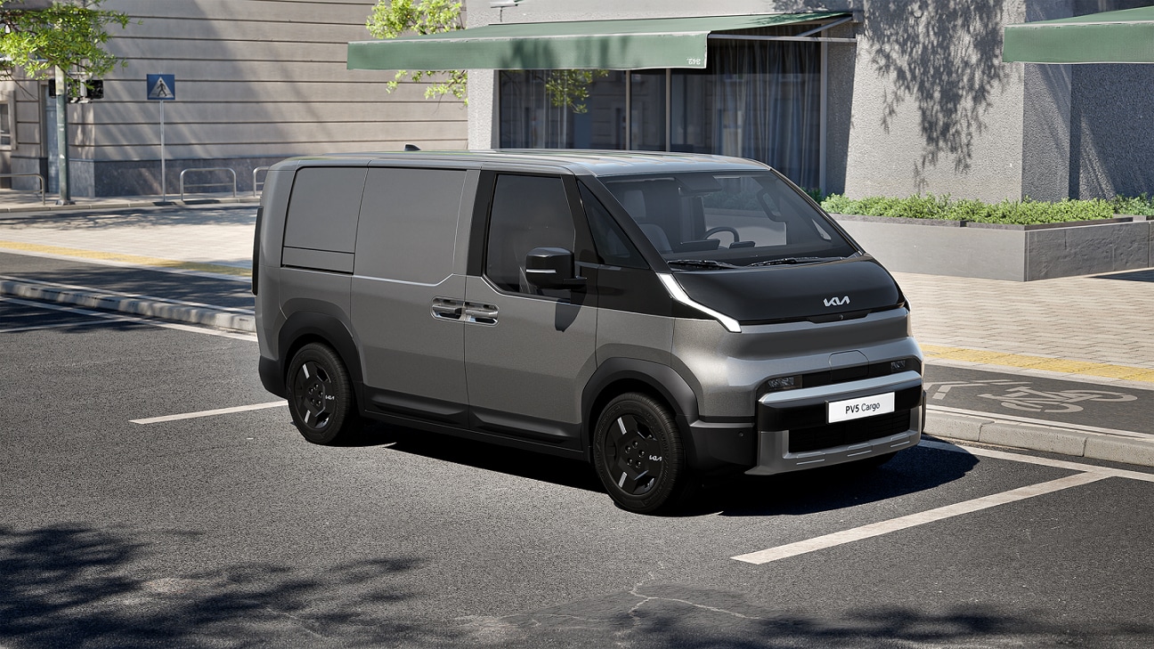 Silberner Kia PV5 Cargo Elektro in der Dreiviertel-Frontansicht, geparkt in einer städtischen Parkbucht am Straßenrand neben einem Gebäude mit grüner Markise