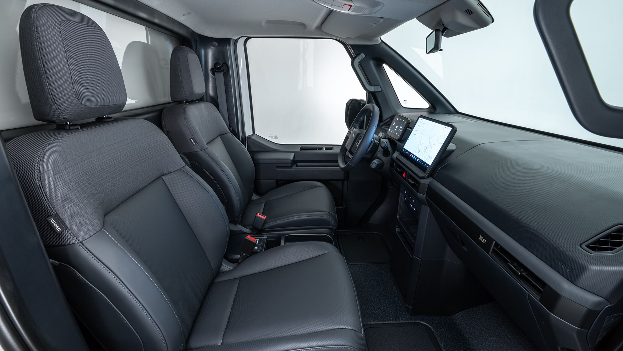 Ein Kia PV5  Chassis-Cab mit zwei dunklen Stoffsitzen, einem aufgesetzten zentralen Infotainment-Display und einem funktionalen, modernen Cockpit-Design