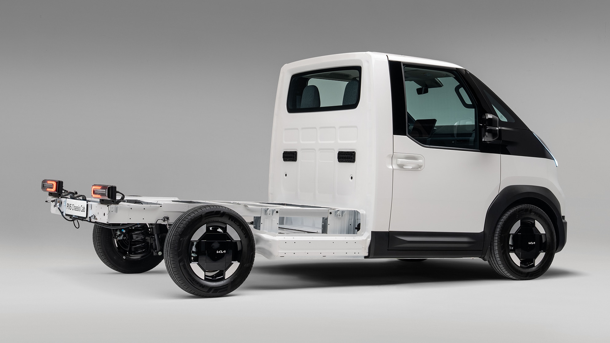 Ein weißer Kia PV5 Chassis Cab Elektro-Transporter in der Heck-Seitenansicht, freigestellt vor einem neutralen grauen Hintergrund